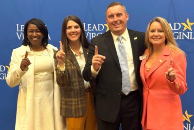 Four people pose for a photo holding up their pointer fingers in front of a blue Lead Virginia backdrop.