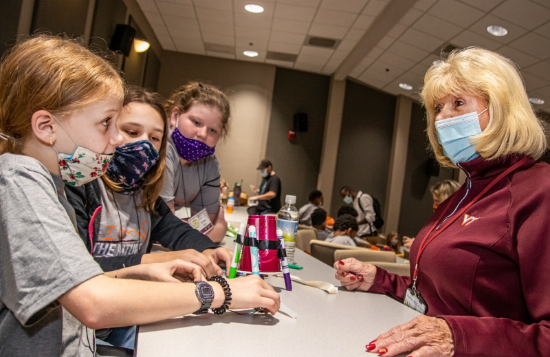 In a large lecture hall, three girls talk to Penny McCallum as a motorized device made from markers and a plastic cup sits between them.